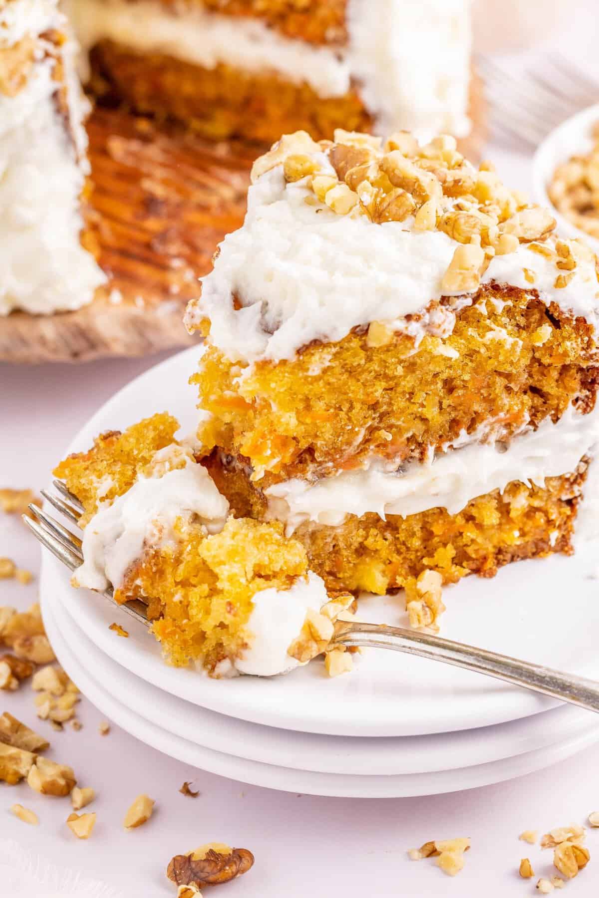 Slice of carrot cake on white stacked plates with fork taking a bite out. 