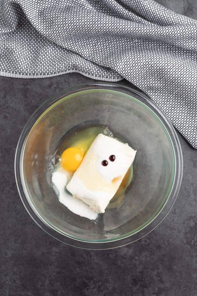 One block of cream cheese, egg, sugar, and red food dye in a large glass bowl. 