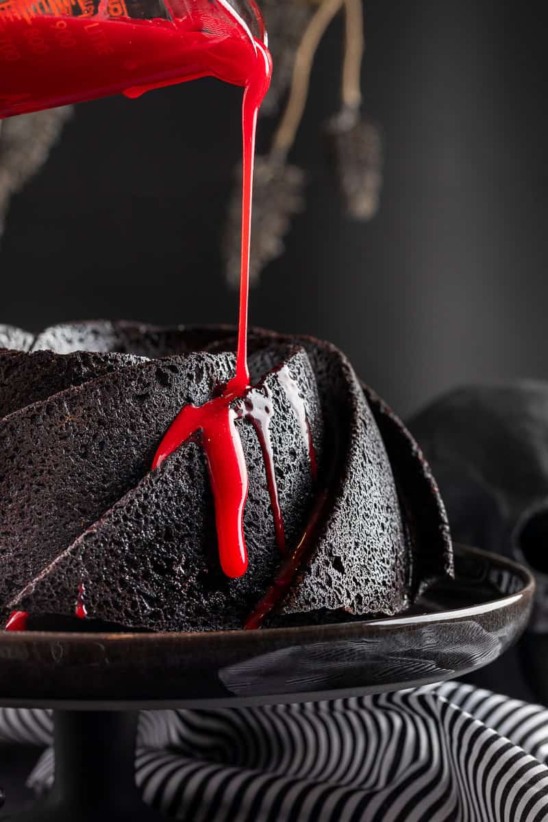 Black bundt cake on a black stand with hand pouring the red ganache over the cake. 