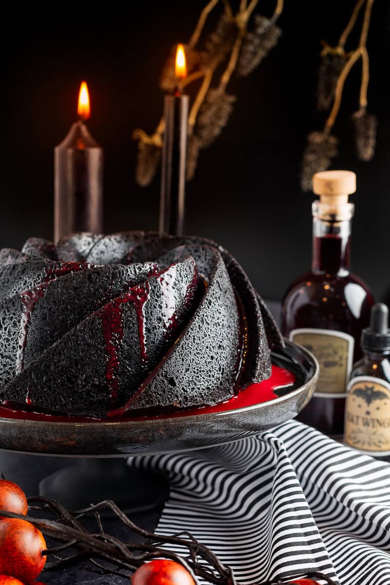 Black bundt cake with red ganache on black cake stand and potion bottles in background. 