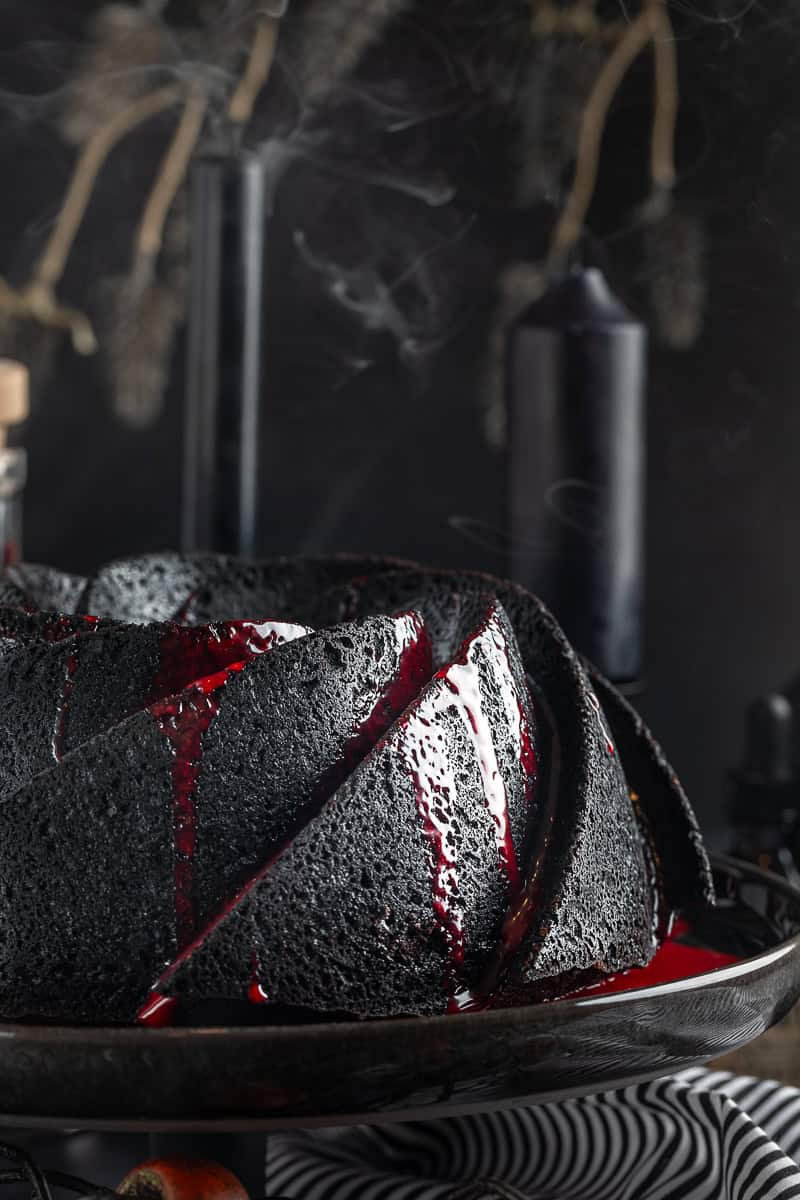 Black bundt cake with red ganache drizzle on a black cake plate and black background. 