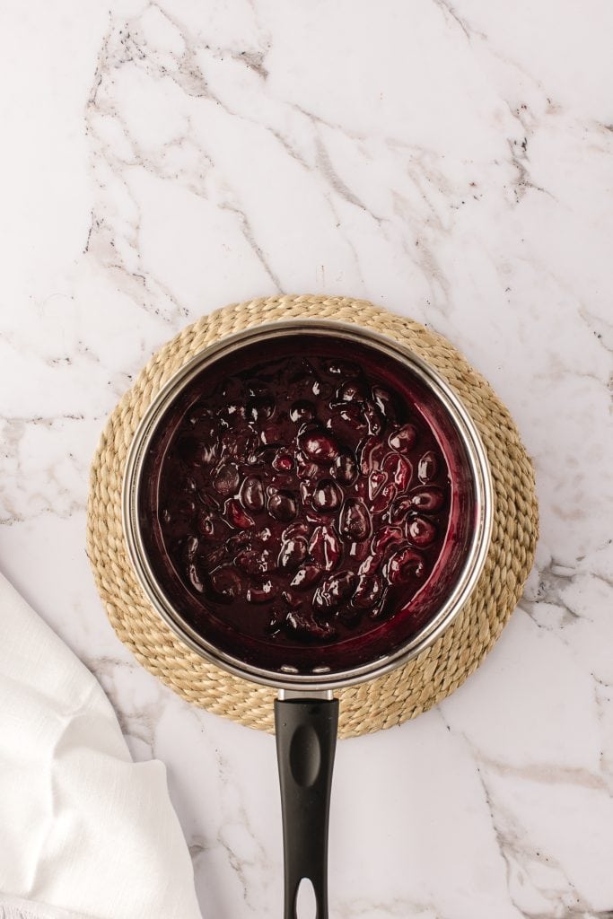 Cooked cherry compote in a small round pan on white marble backdrop. 