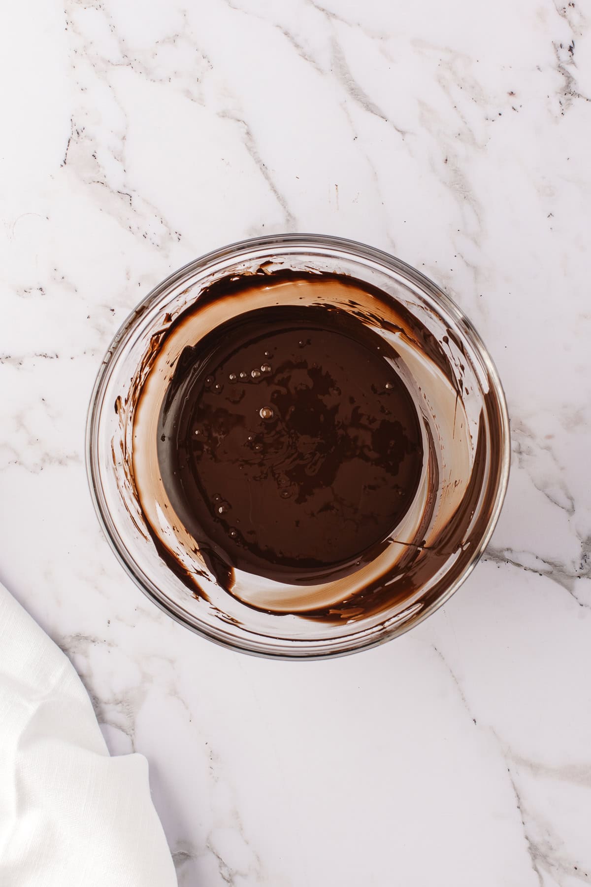 Melted chocolate in a large glass bowl on a white marble backdrop. 