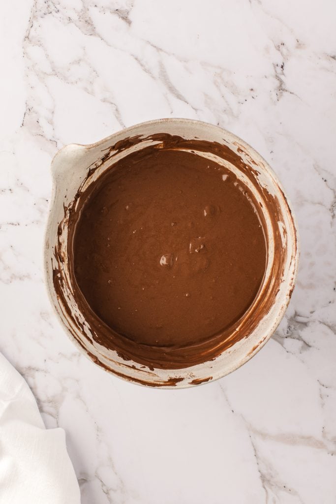 Chocolate cheesecake filling in a large white mixing bowl on a white marble background. 